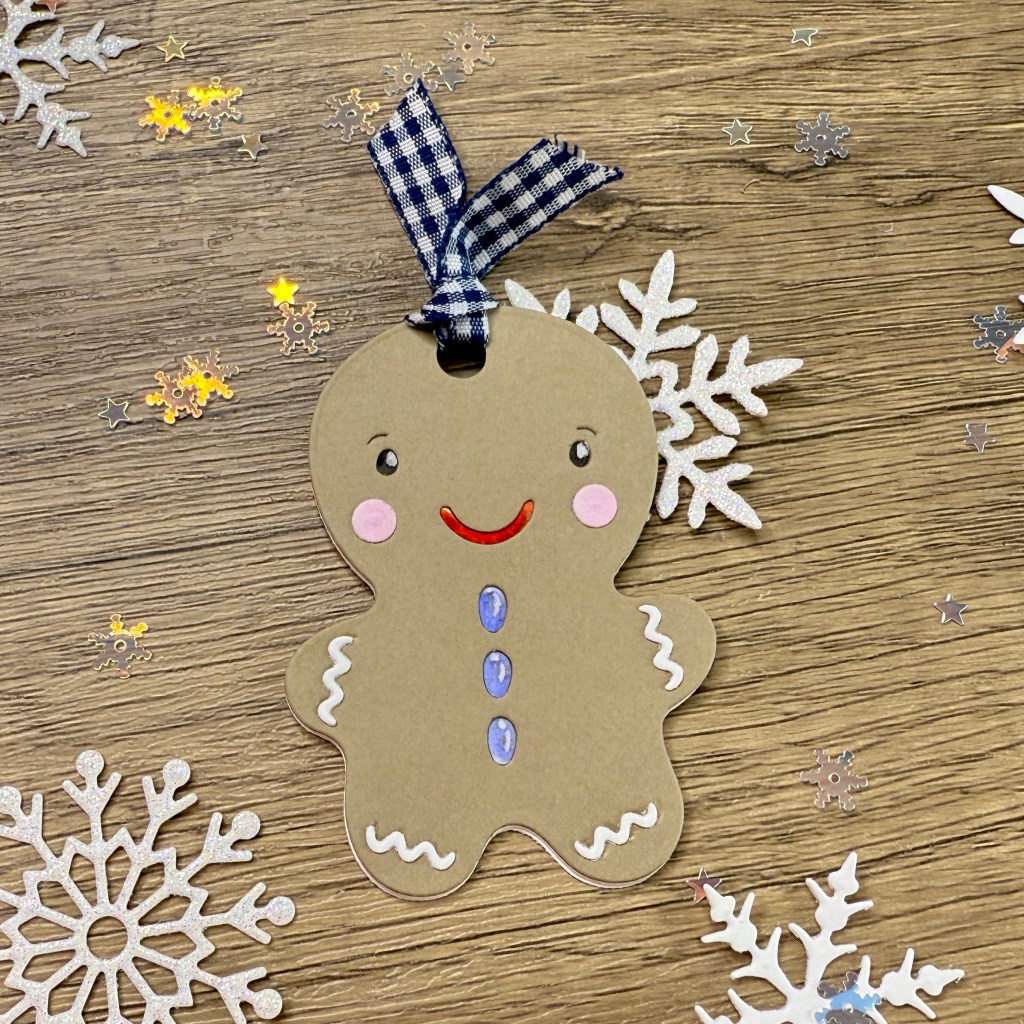 A gingerbread man gift tag with painted facial features, blue buttons, and white icing details, hanging from a blue and white gingham ribbon, surrounded by silver snowflake confetti on a wooden surface.