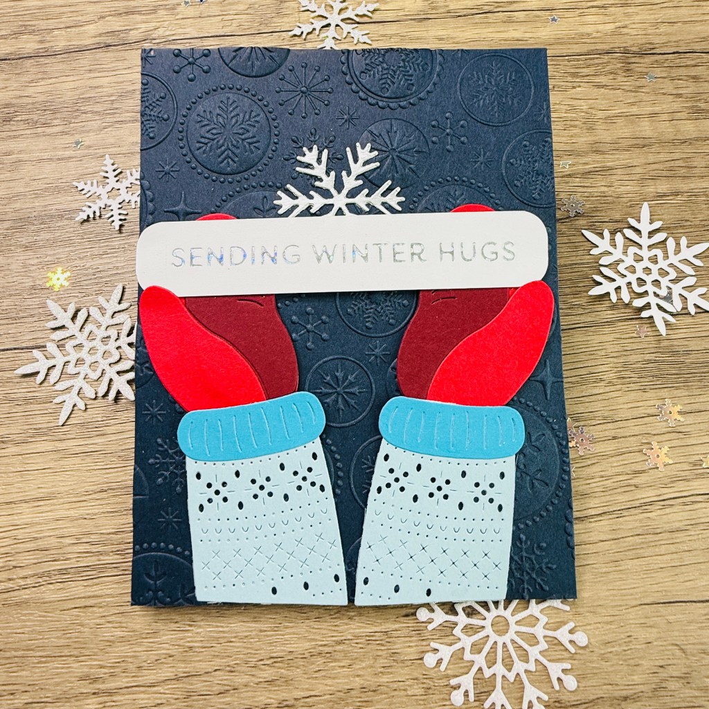 A handmade card featuring red mittens with blue cuffs and a message reading 'SENDING WINTER HUGS' placed in the center, set against an embossed dark blue background with snowflake designs and scattered snowflake embellishments.