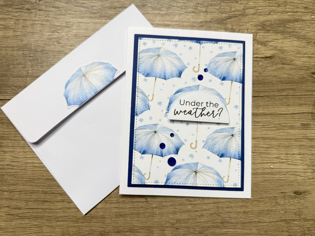 Handmade card featuring blue umbrella patterns with the sentiment 'Under the weather?', accompanied by a matching envelope.