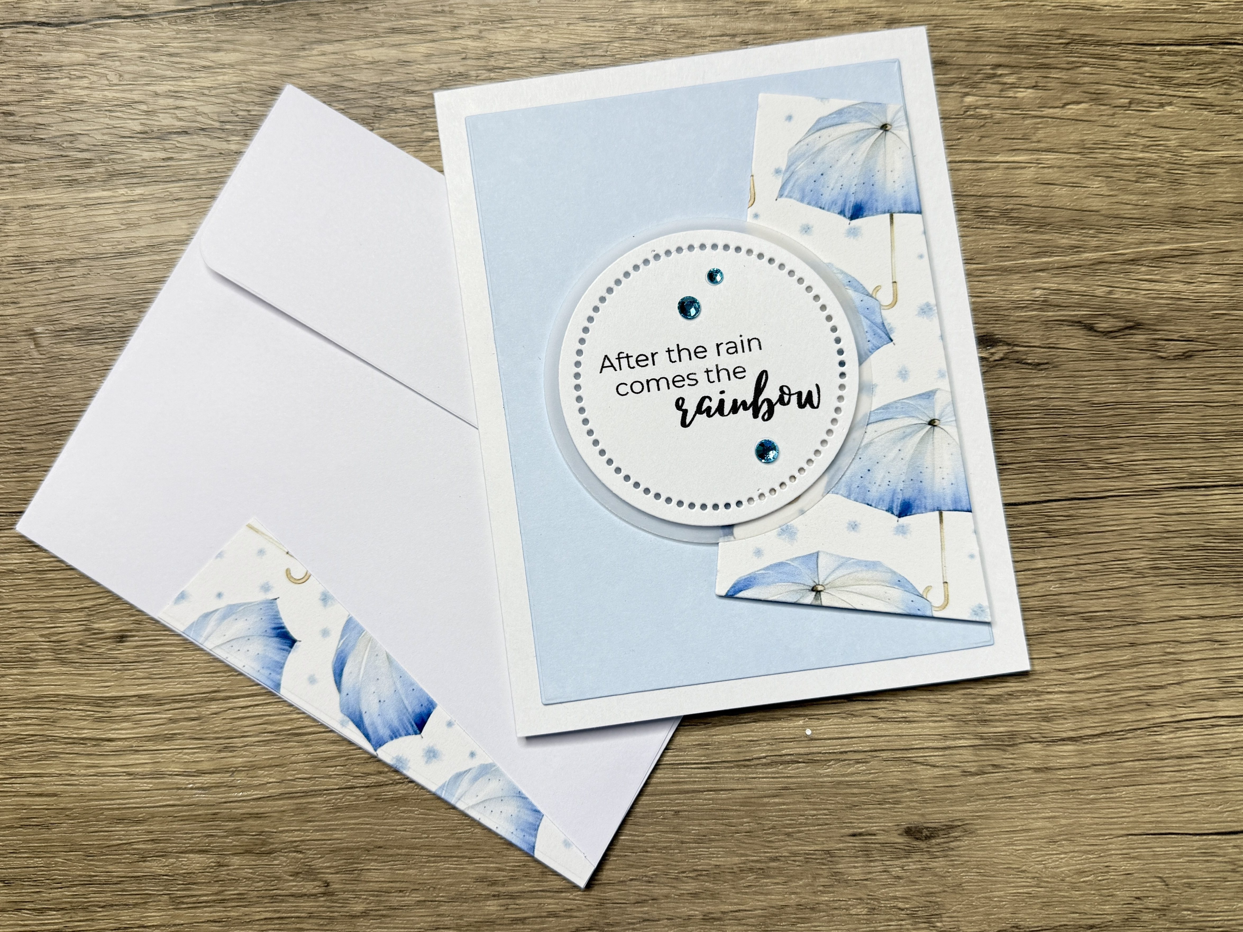 A handmade card featuring a blue background with an umbrella design, showcasing the sentiment 'After the rain comes the rainbow' in stylish typography, accompanied by decorative gems. The card is paired with a coordinating envelope featuring a similar umbrella motif.