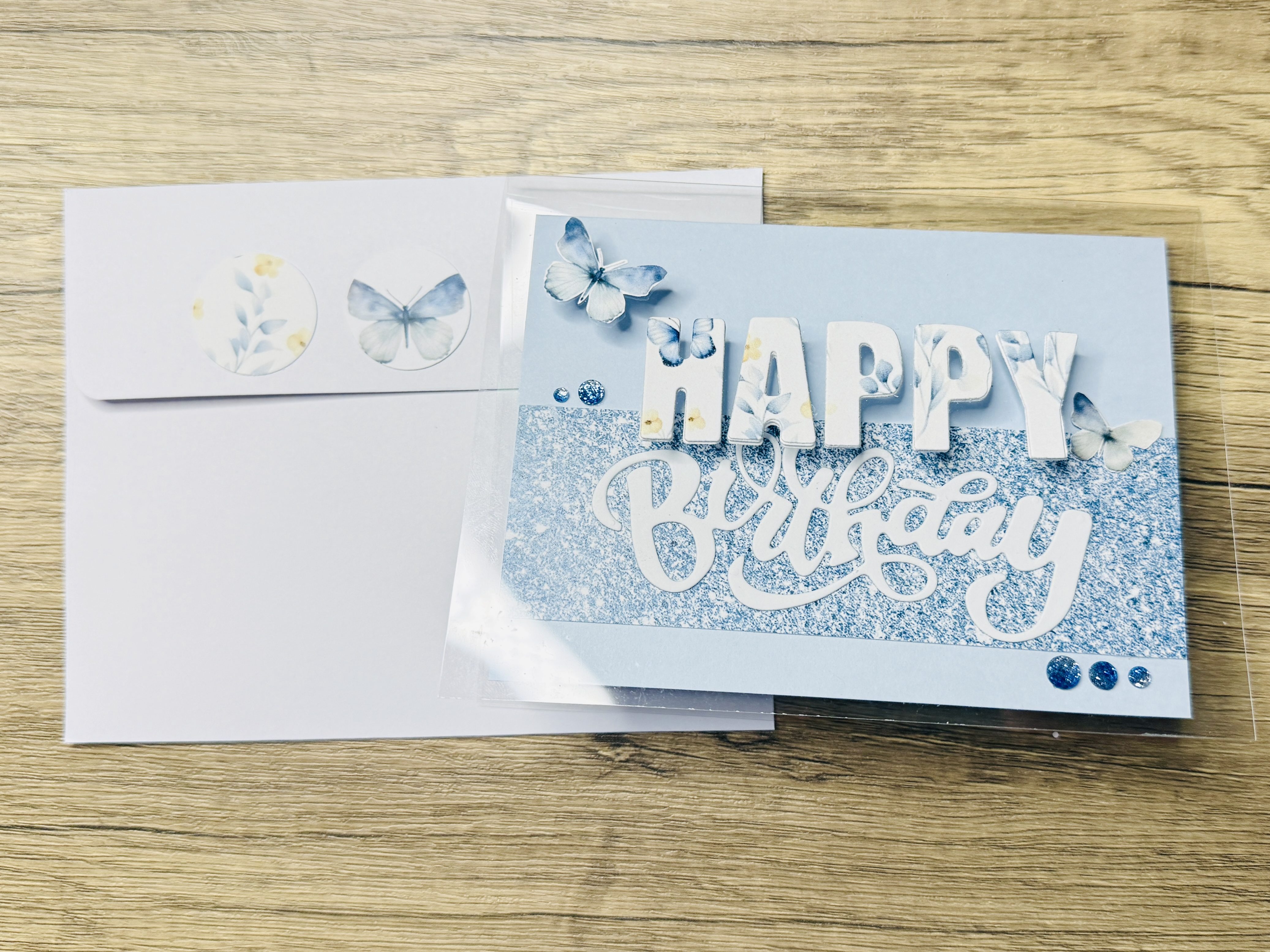 A handcrafted birthday card featuring an acetate card base with layered blue paper with the text 'Happy Birthday' in white, embellished with butterflies and sparkling gems, placed next to a coordinating envelope adorned with decorative circular stickers.