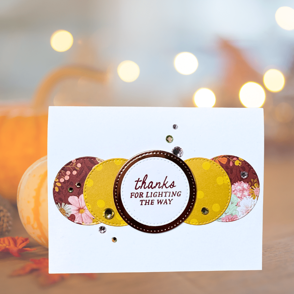 A handmade card featuring a circular design with patterned circles in autumn colors, adorned with sequins and the sentiment 'thanks for lighting the way' in the center.