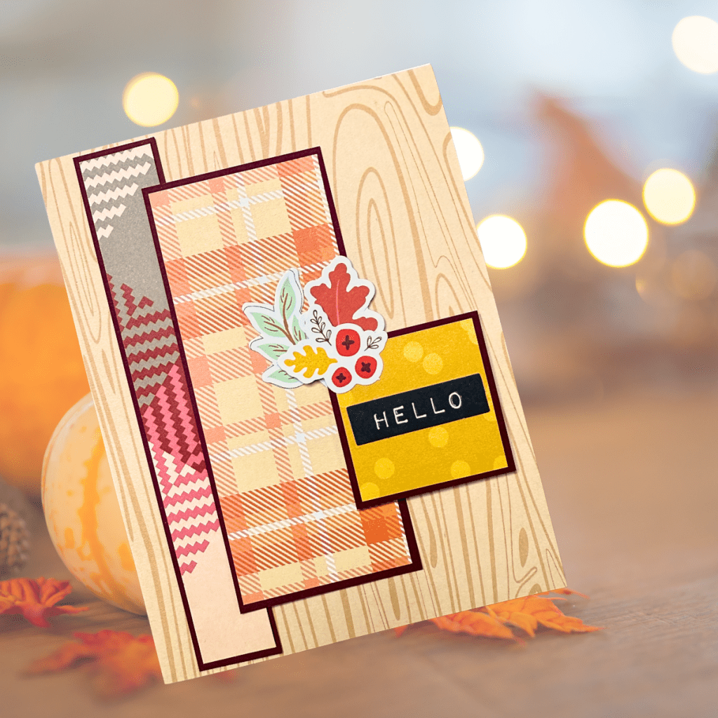 A handmade greeting card featuring warm autumn colors, patterned paper with a wood grain design, and embellishments including leaves and a black 'HELLO' sentiment.