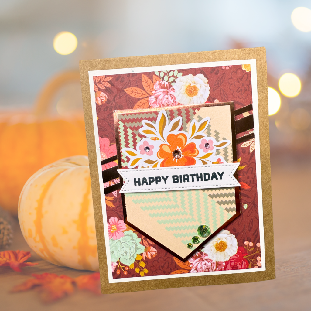 A handmade birthday card featuring floral patterned paper and a central banner that reads 'HAPPY BIRTHDAY', adorned with decorative gems.