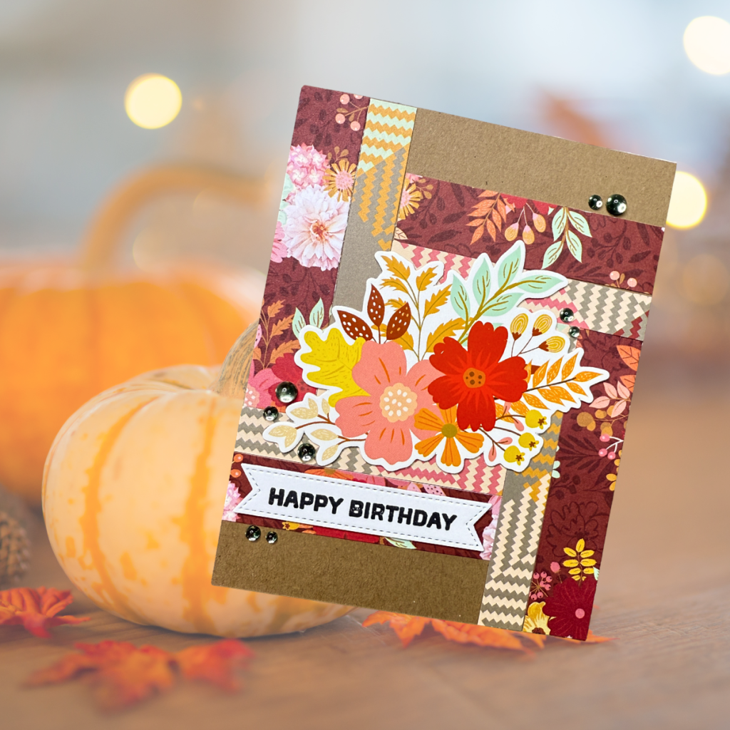 A handcrafted birthday card featuring a collage of patterned paper in warm autumn colors, adorned with floral die-cuts and a 'HAPPY BIRTHDAY' sentiment placed on a banner.