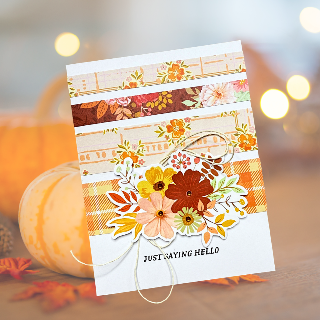 A greeting card featuring patterned paper strips in autumn colors, adorned with a floral arrangement and twine, with the text 'JUST SAYING HELLO' at the bottom.
