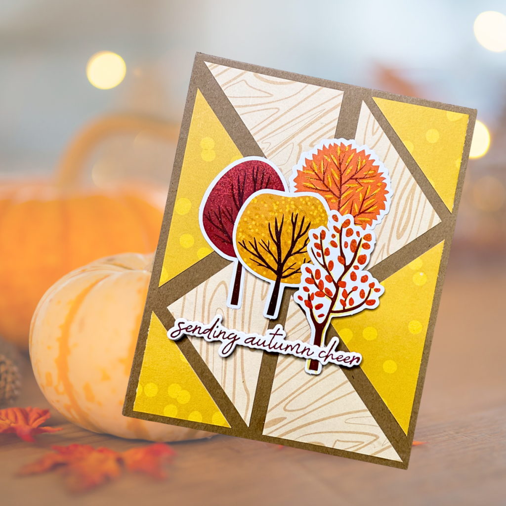 A handcrafted greeting card featuring colorful autumn trees, including red, orange, and yellow foliage, layered on a geometric background. The text 'sending autumn cheer' is prominently displayed.