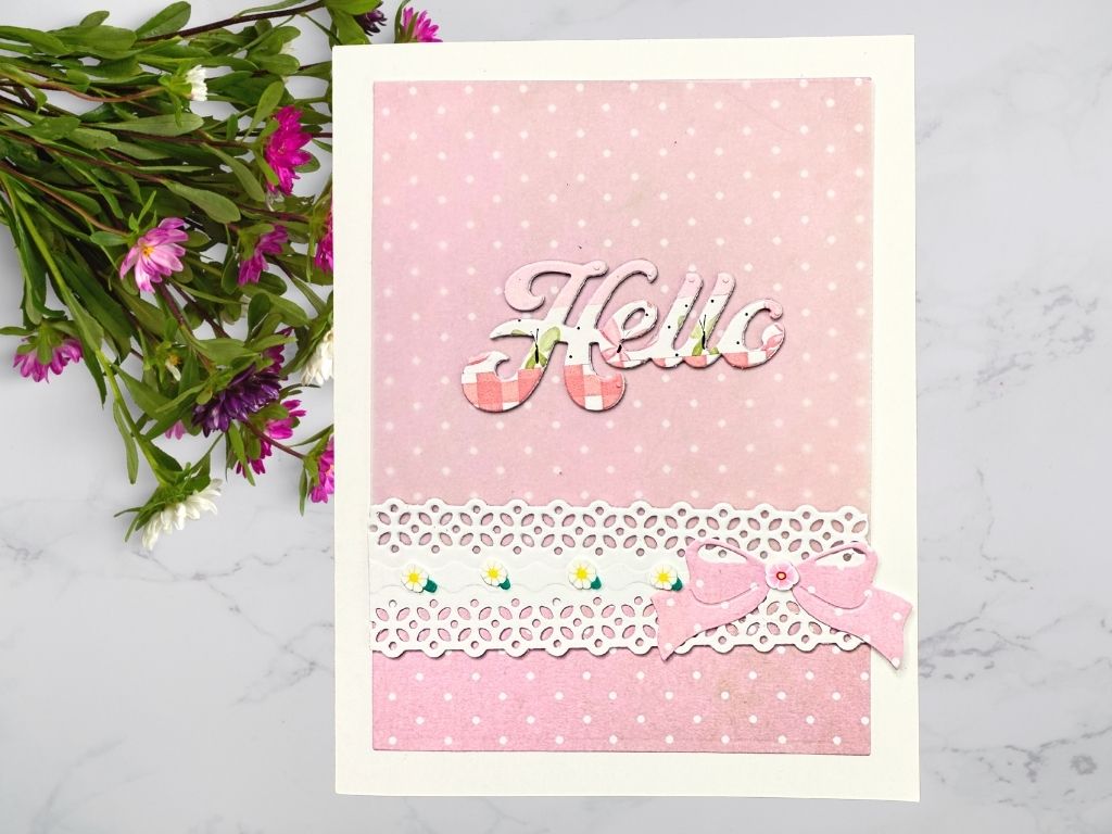 A handmade card featuring a pink and white polka dot background with a layered 'Hello' sentiment. The card has a decorative lace border at the bottom and is embellished with a pink bow and small floral accents.