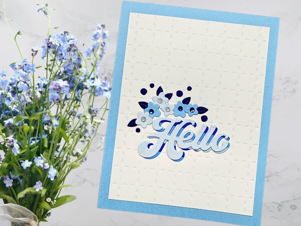 A handmade card featuring a layered blue and white 'Hello' sentiment adorned with small floral embellishments, set against a textured background and a light blue card base.