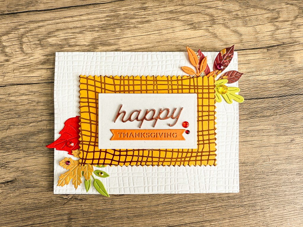 A handmade Thanksgiving card featuring a yellow plaid frame around the words 'happy THANKSGIVING,' decorated with colorful autumn leaves and gems on a textured white background.
