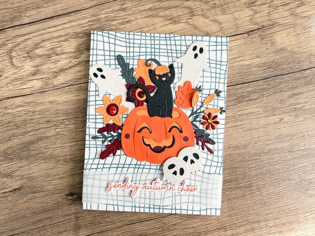 A vibrant autumn-themed card featuring a smiling pumpkin, a playful black cat, ghost decorations, and colorful flowers, with the text 'sending autumn cheer' at the bottom.