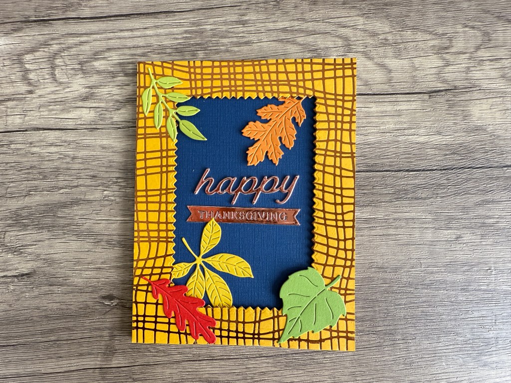 A handmade card featuring a blue center with the text 'happy THANKSGIVING' in copper foil, surrounded by a yellow grid pattern and decorated with various colorful leaf embellishments.