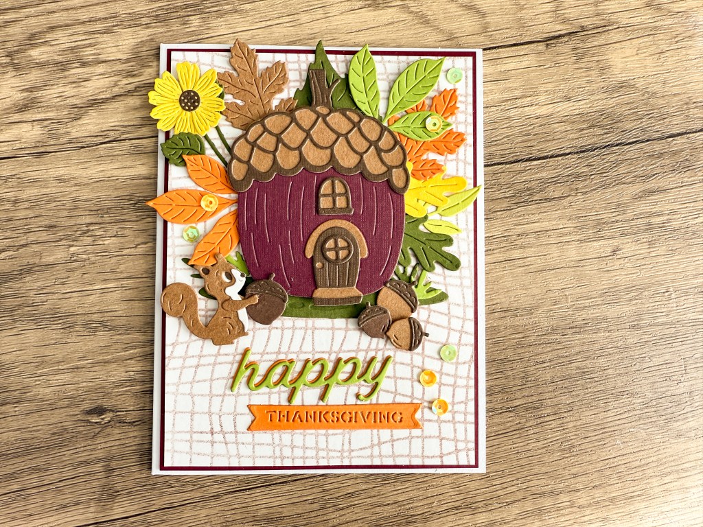 A festive Thanksgiving card featuring a whimsical acorn house surrounded by colorful fall leaves, flowers, and a playful squirrel holding an acorn.