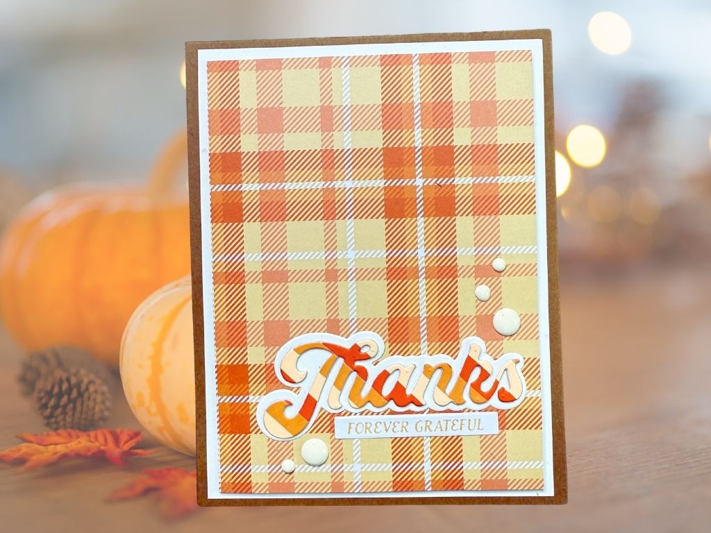 A handmade card featuring a plaid design in warm autumn colors, with the word 'Thanks' in a bold, layered font and the phrase 'FOREVER GRATEFUL' underneath. The card is accented with small, circular embellishments and has a brown card base.