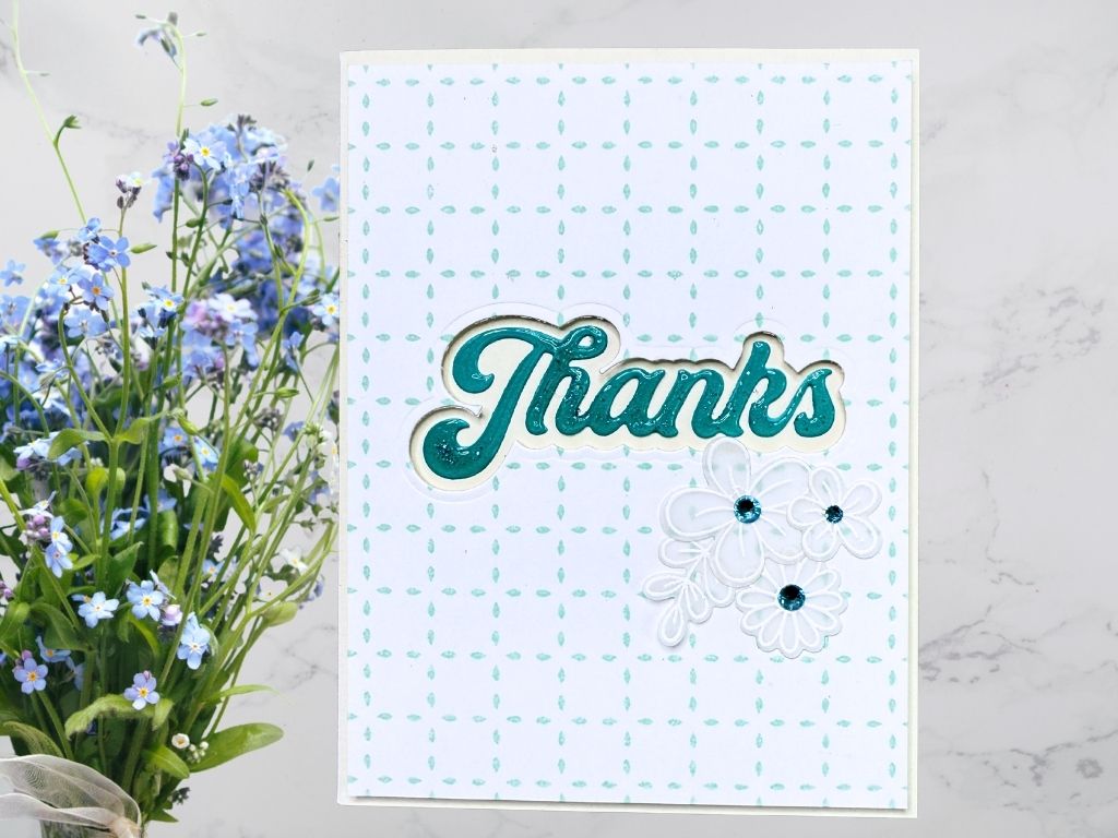 A handmade card featuring the word 'Thanks' in a glossy teal finish, surrounded by white floral embellishments and a white background with light blue BetterPress pattern.