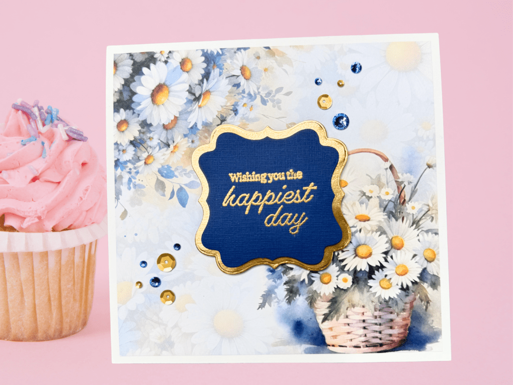 A handmade greeting card with a floral design featuring daisies, a gold-embossed sentiment that reads 'Wishing you the happiest day,' and decorative sequins, displayed next to a pink cupcake.
