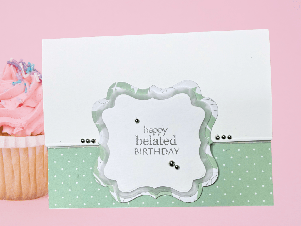 A handmade card featuring a layered design with a scalloped label cutout, embossed text reading 'happy belated BIRTHDAY,' and decorative elements like silver pearls and a green polka dot background. 