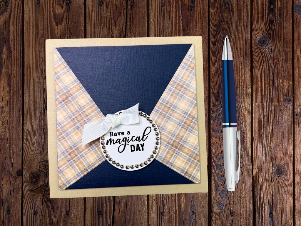 A handmade card featuring a plaid design on the left side, a solid navy blue right panel, and a circular sentiment that reads 'Have a magical day' with decorative silver embellishments. The card is presented on a wooden surface alongside a blue and silver pen.