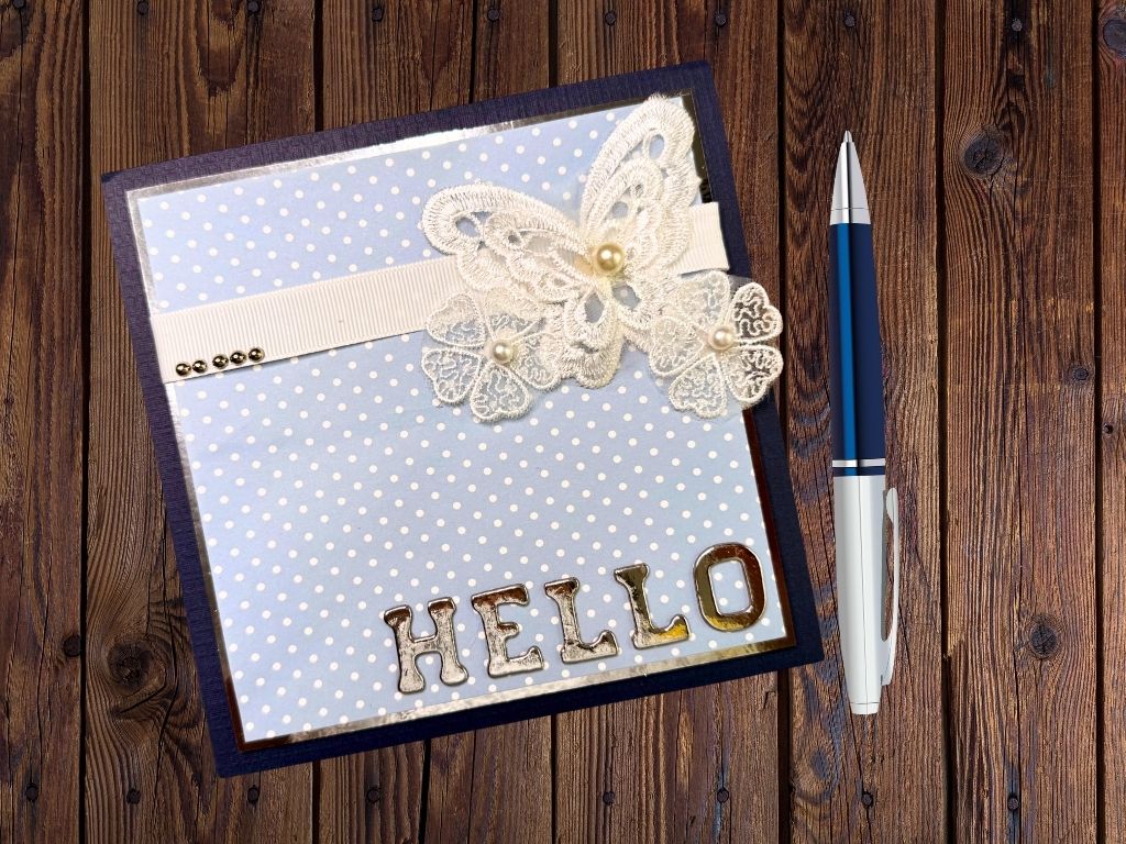 A handmade card with a blue polka dot background featuring a lace butterfly, flower embellishments, and the word 'HELLO' in shiny letters. The card is placed on a wooden surface next to a blue pen.