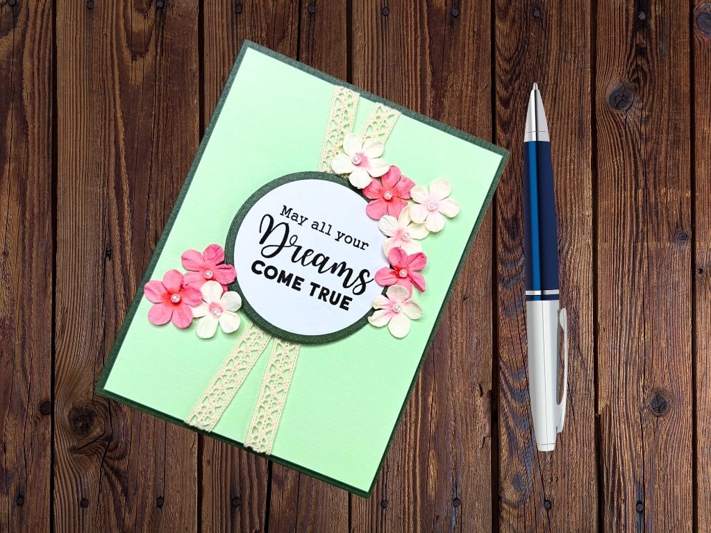 A handmade card featuring a light green background with a circular sentiment that reads 'May all your Dreams COME TRUE,' surrounded by pink and white paper flowers and lace ribbon decorations, placed on a wooden surface next to a pen.