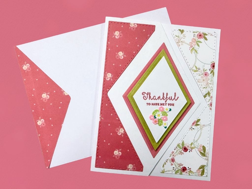 An asymmetrical fold thank-you card featuring a layered diamond design with a floral background, the text 'Thankful to have met you' centered on a white panel, surrounded by pink and green accents.