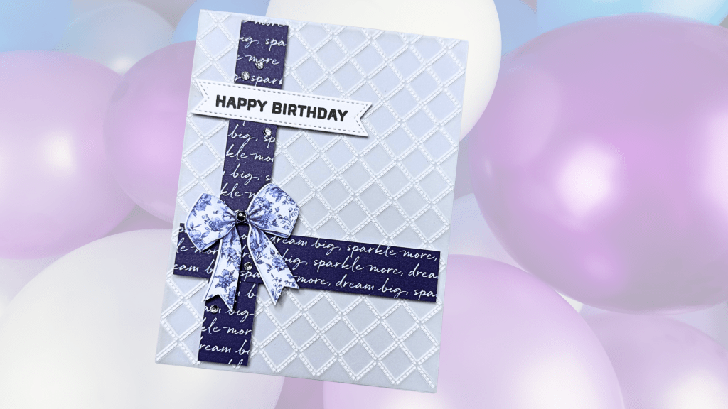 A birthday card featuring a light blue background with a diamond pattern, adorned with a purple ribbon and a floral bow. The card includes the text 'HAPPY BIRTHDAY' in a white banner at the top, with purple balloons blurred in the background.