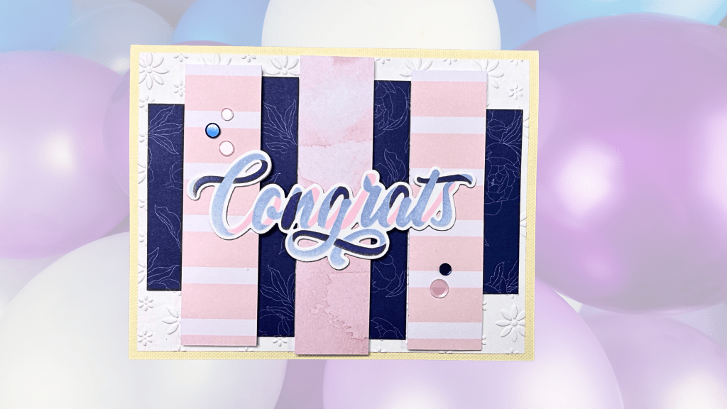 A handmade card featuring the word 'Congrats' in cursive, adorned with patterned paper stripes in pink and navy, with embellishments like round gems, set against a soft pastel balloon background.