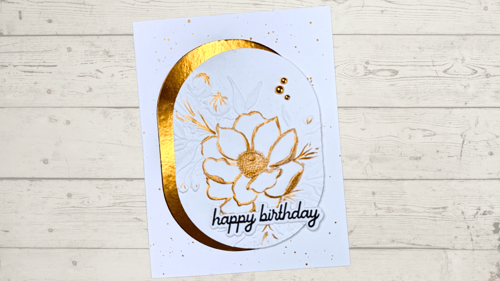 A handmade birthday card featuring an embossed floral design in white with gold metallic accents, framed by a shiny gold oval border and adorned with decorative gems.