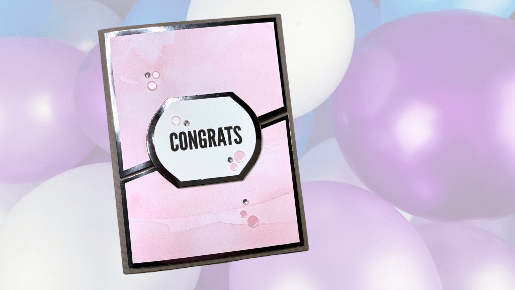 A handmade card featuring the word 'CONGRATS' on a pink watercolor background, accented with small embellishments, set against a backdrop of pastel-colored balloons.