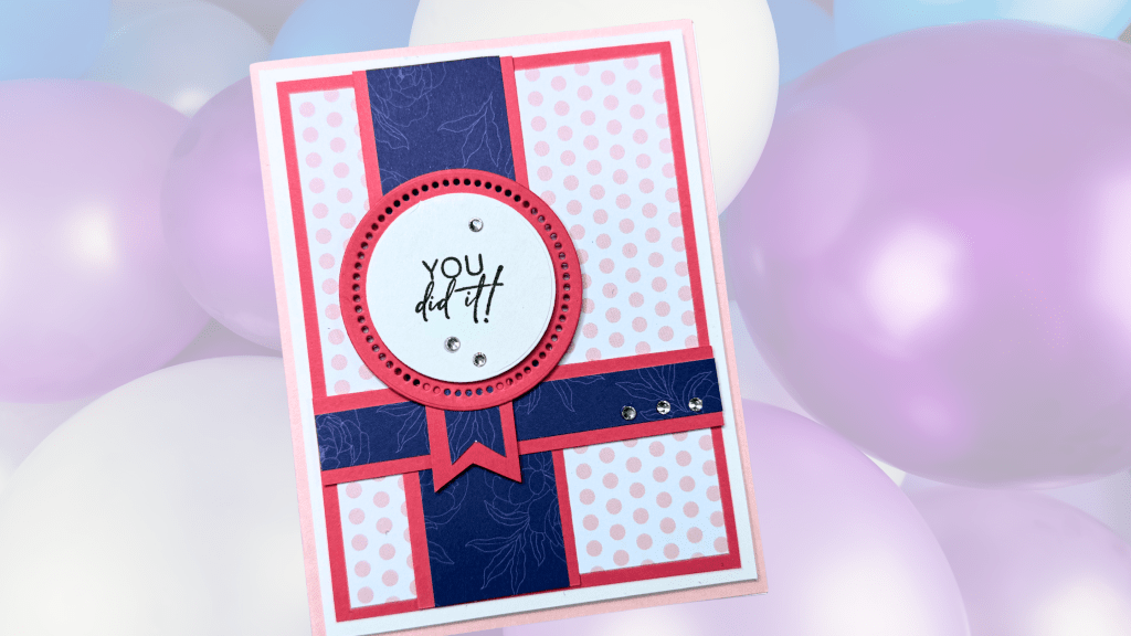 A handcrafted card featuring a circular label that says 'YOU did it!' surrounded by a peppy design of pink and navy blue patterned paper, set against a soft background of pastel balloons.