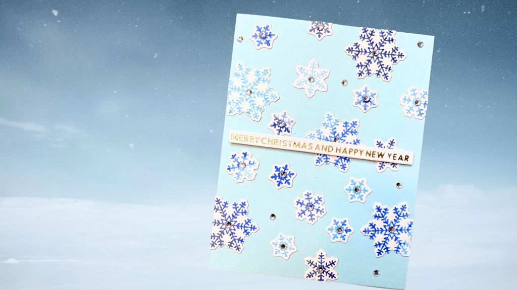A festive blue card adorned with various snowflake designs in white and shades of blue, featuring a gold sentiment that reads 'Merry Christmas and Happy New Year'. Sparkling gems are placed at the centers of some snowflakes, creating a cheerful holiday theme.