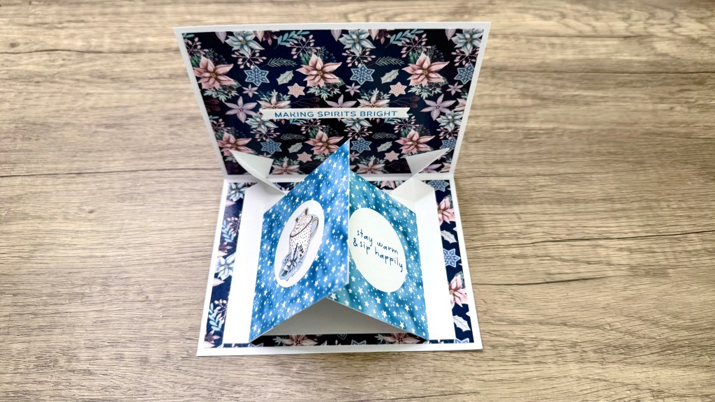 A vibrant double pop-up card featuring floral patterns and a blue starry insert with a festive message, displayed with the card open on a wooden surface.