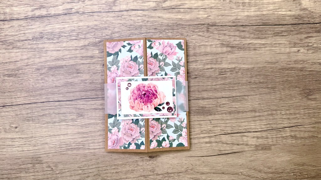 A handmade explosion fold pop-up card featuring floral patterned paper with a central embellishment, displayed on a wooden surface.