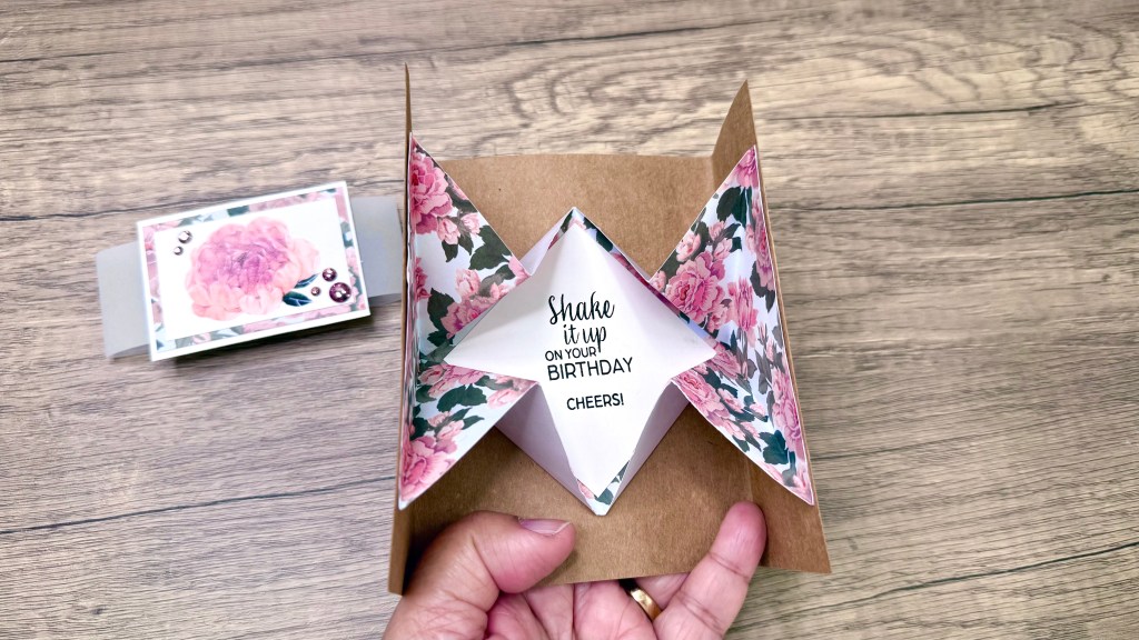 An open explosion card featuring a pop-up design with floral paper and a birthday message. The card is held in a hand, showcasing the intricate folds and embellishments.