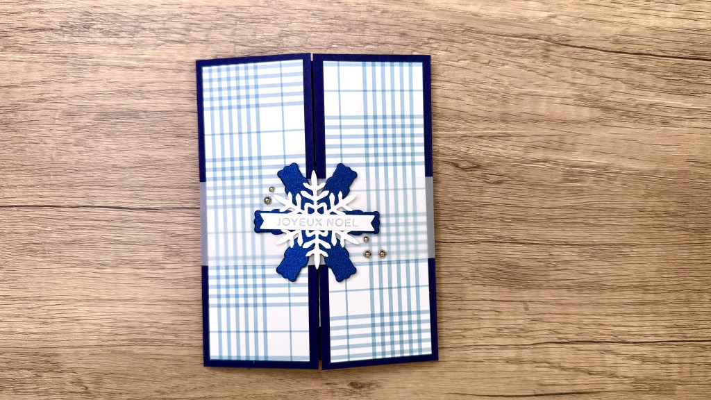 A handmade explosion fold pop-up card with blue plaid patterned paper, a white snowflake, and the text 'Joyeux Noel' positioned in the center.