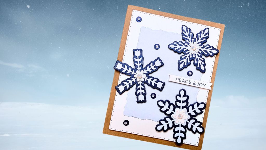 Layered snowflake holiday card featuring intricate die cuts, rhinestone embellishments, and a sentiment strip reading 'PEACE & JOY' on a textured background.