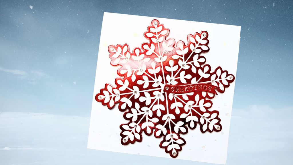 A red and white snowflake card with the word 'GREETINGS' in the center, surrounded by small embellishments. The card is set against a snowy background.