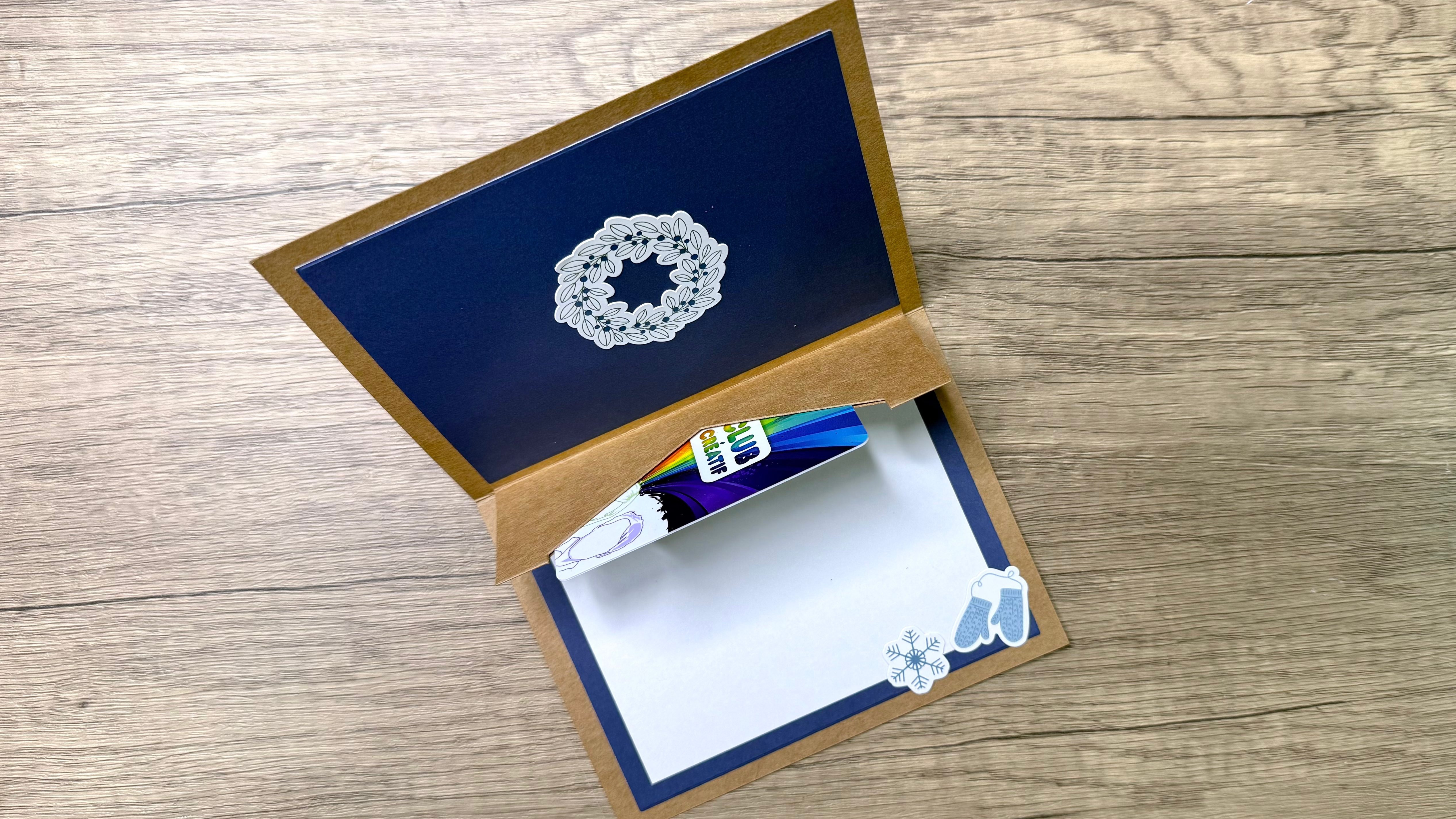 An open holiday card featuring a navy blue panel with a wreath design, showing a gift card holder and some embellishments like snowflake and mitten stickers.