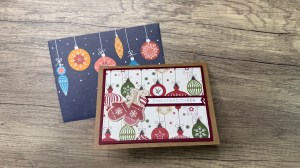 A holiday card featuring colorful ornaments with a clean and modern design, placed next to a blue card adorned with hanging ornaments on a wooden surface.