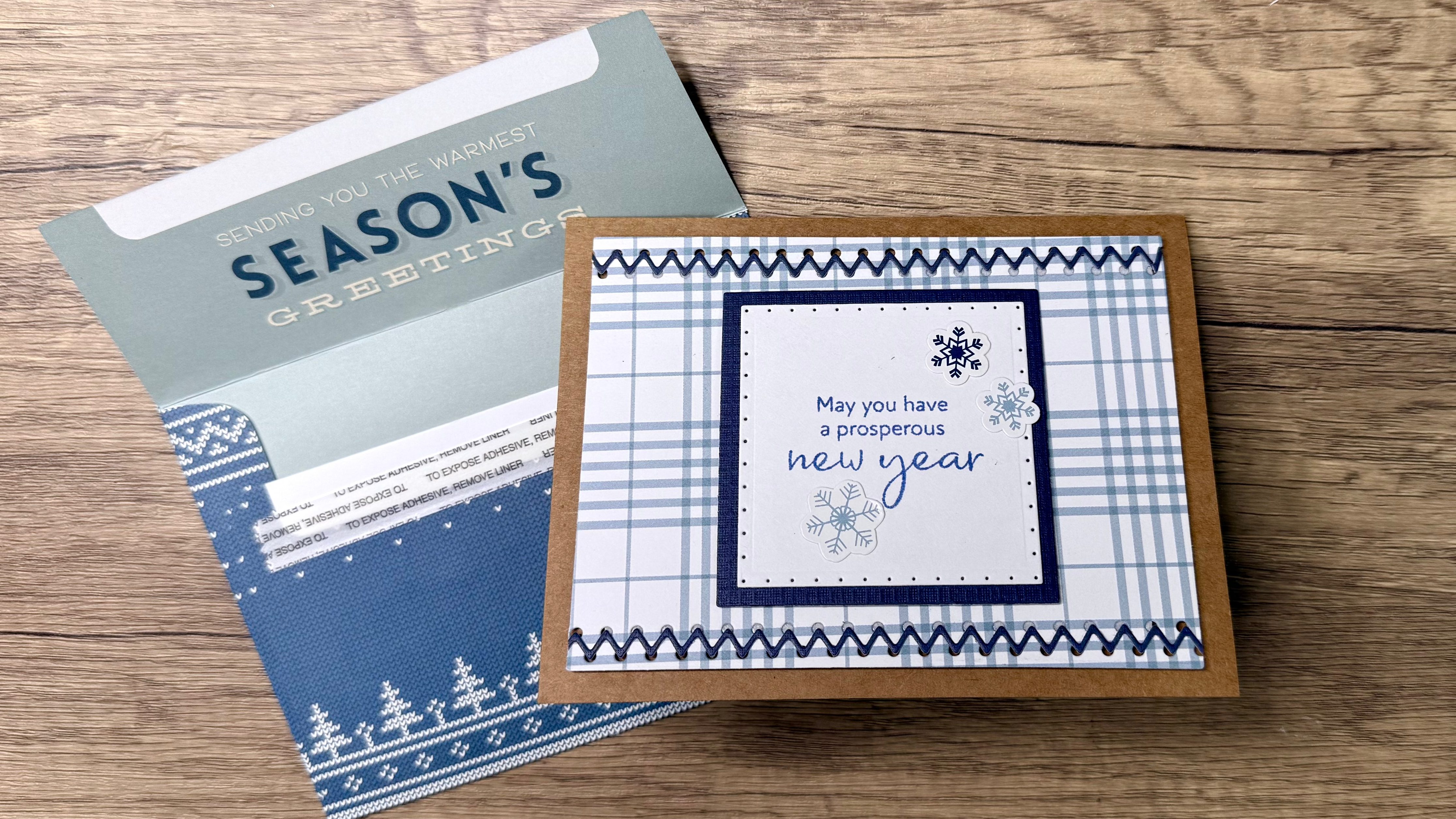 A handmade holiday card featuring a blue and white plaid design with a sentiment that reads 'May you have a prosperous new year,' placed on a kraft cardstock base next to a decorative envelope that has a winter theme.