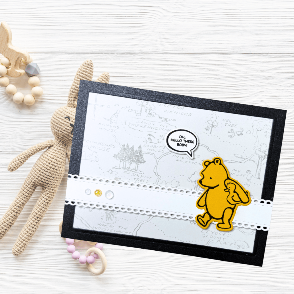 A gender-neutral baby card featuring a playful yellow bear character with a speech bubble saying 'Oh, hello there baby', set against a subtle grey background, framed by a black cardstock border, and accompanied by a textured white decorative band.