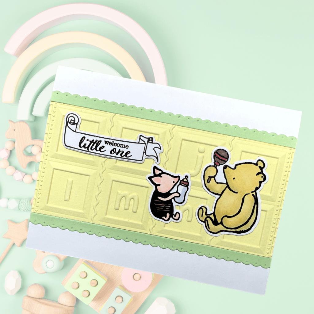 A cheerful, gender-neutral baby card featuring embossed yellow cardstock with playful characters, including a bear and a pig, holding a bottle and a snack, accompanied by a banner that reads 'welcome little one'. The design is accented with a soft green scalloped border.