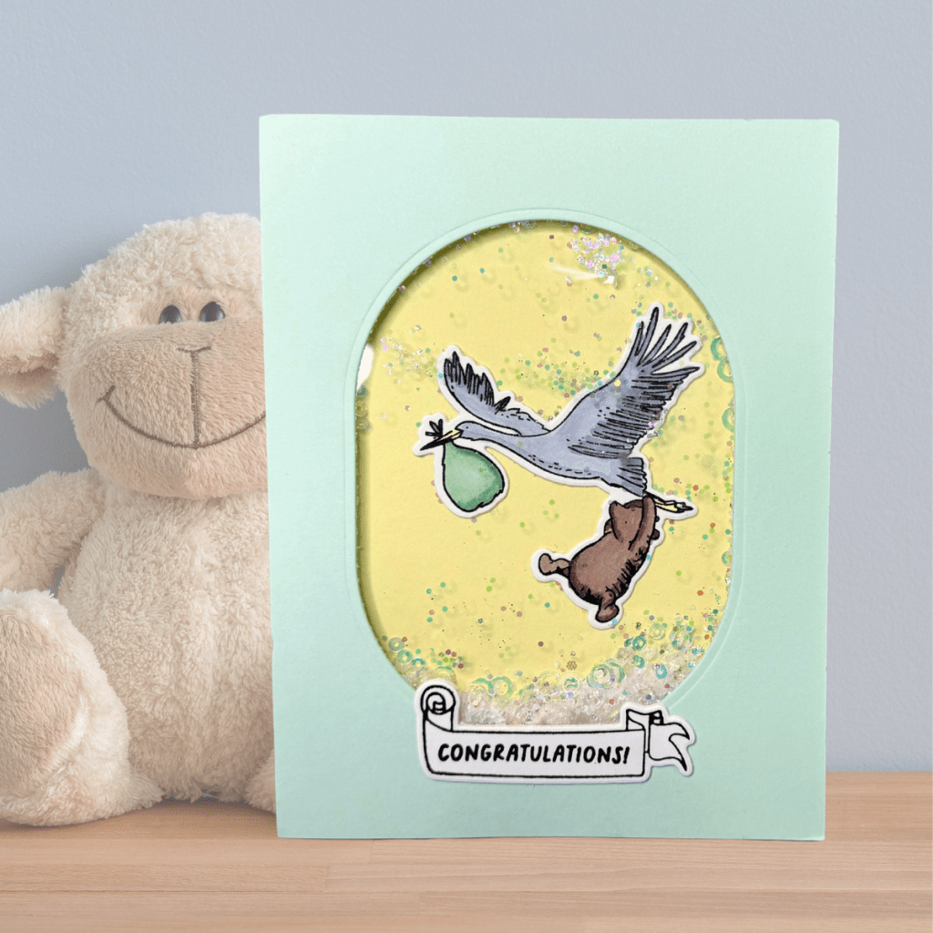 A gender-neutral baby card featuring a stork carrying a bundle, surrounded by a shaker window with yellow and green colors, and a 'Congratulations!' banner at the bottom.