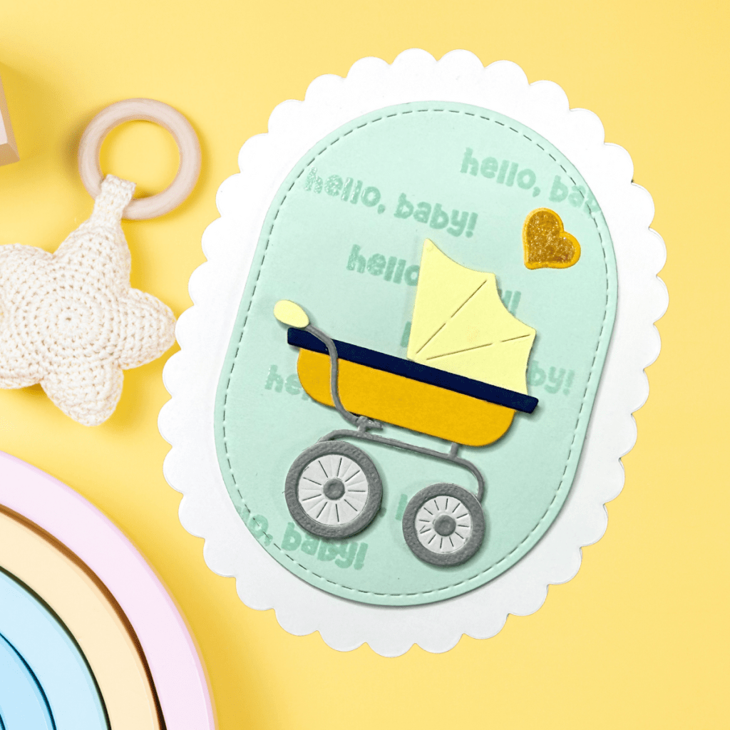A shaped baby card featuring a vintage-style pram in yellow and navy, with a mint green background that includes 'hello, baby!' text and a small golden heart.