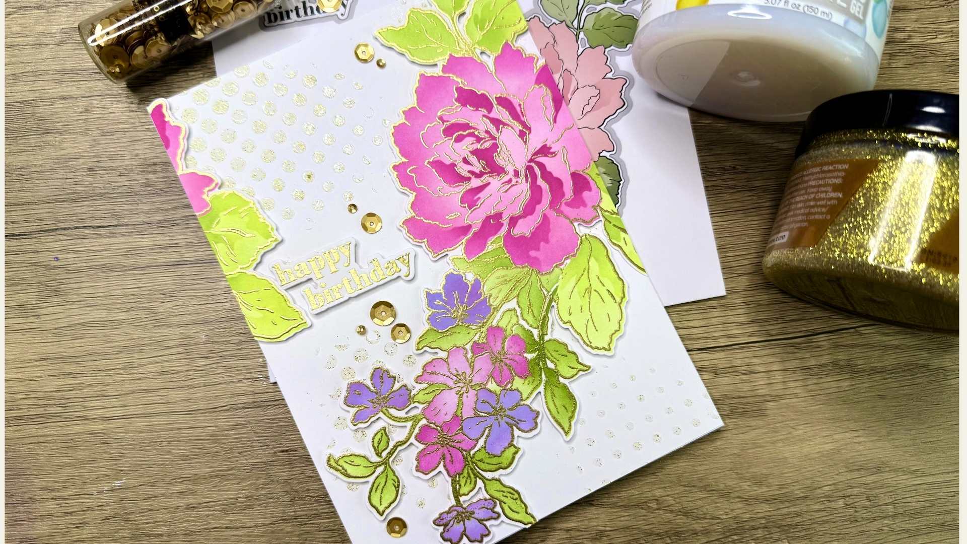 Handmade birthday card made with Altenew products with pink and purple flowers, green leaves, and gold heat embossing and embellishments on a wooden surface