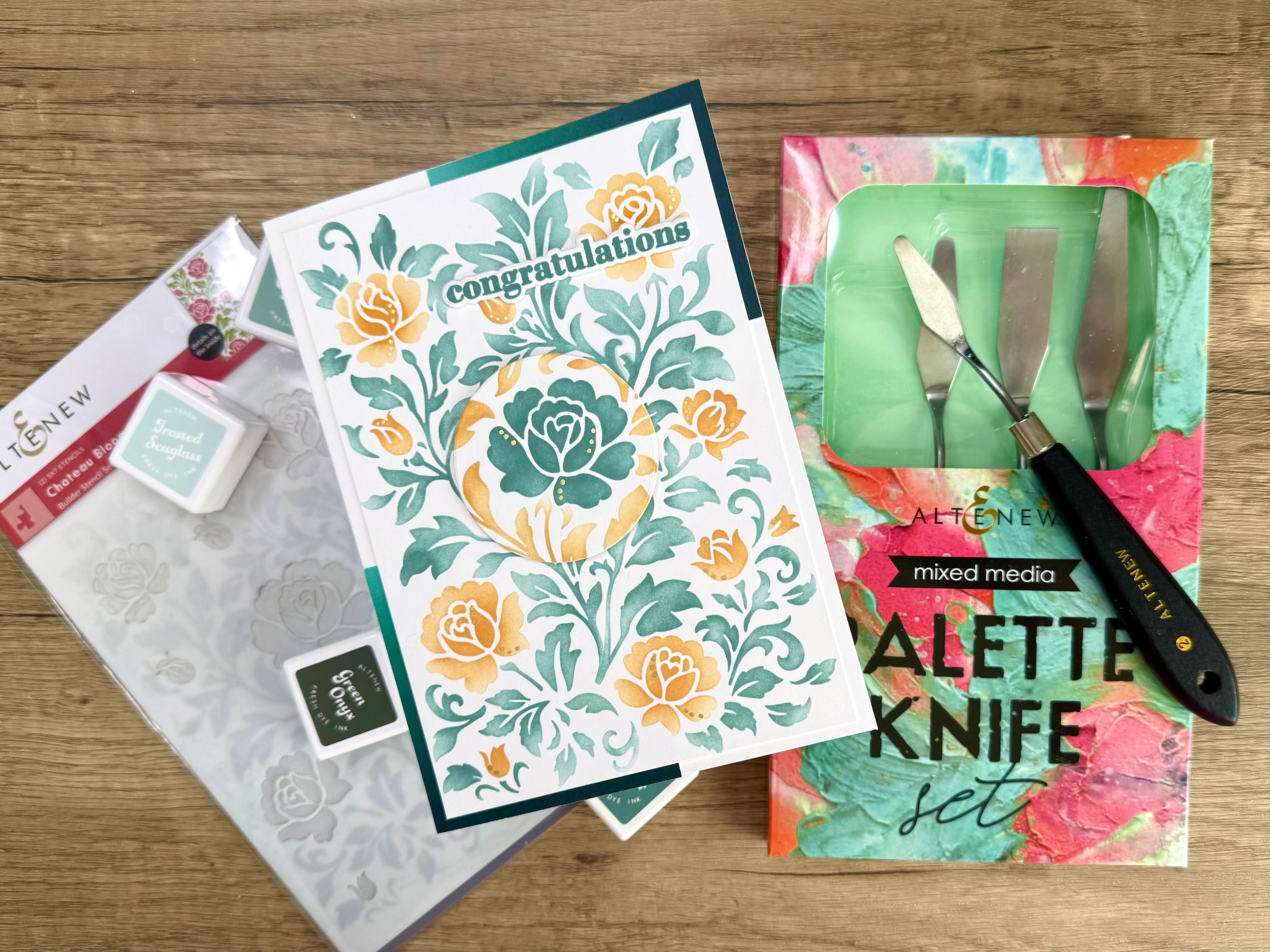 IMG_2399 Congratulations card with floral design in teal and orange, art stencils, ink pads in Frosted Seaglass and Green Olive, and a mixed media palette knife set on a wooden surface